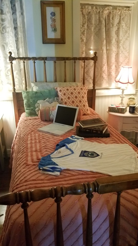 A narrow twin bed with a pink comforter. Draped over the bed is v neck chilton baseball tee. Also laying on the bed some classic literary texts, an old macbook, and fluffy pillows.